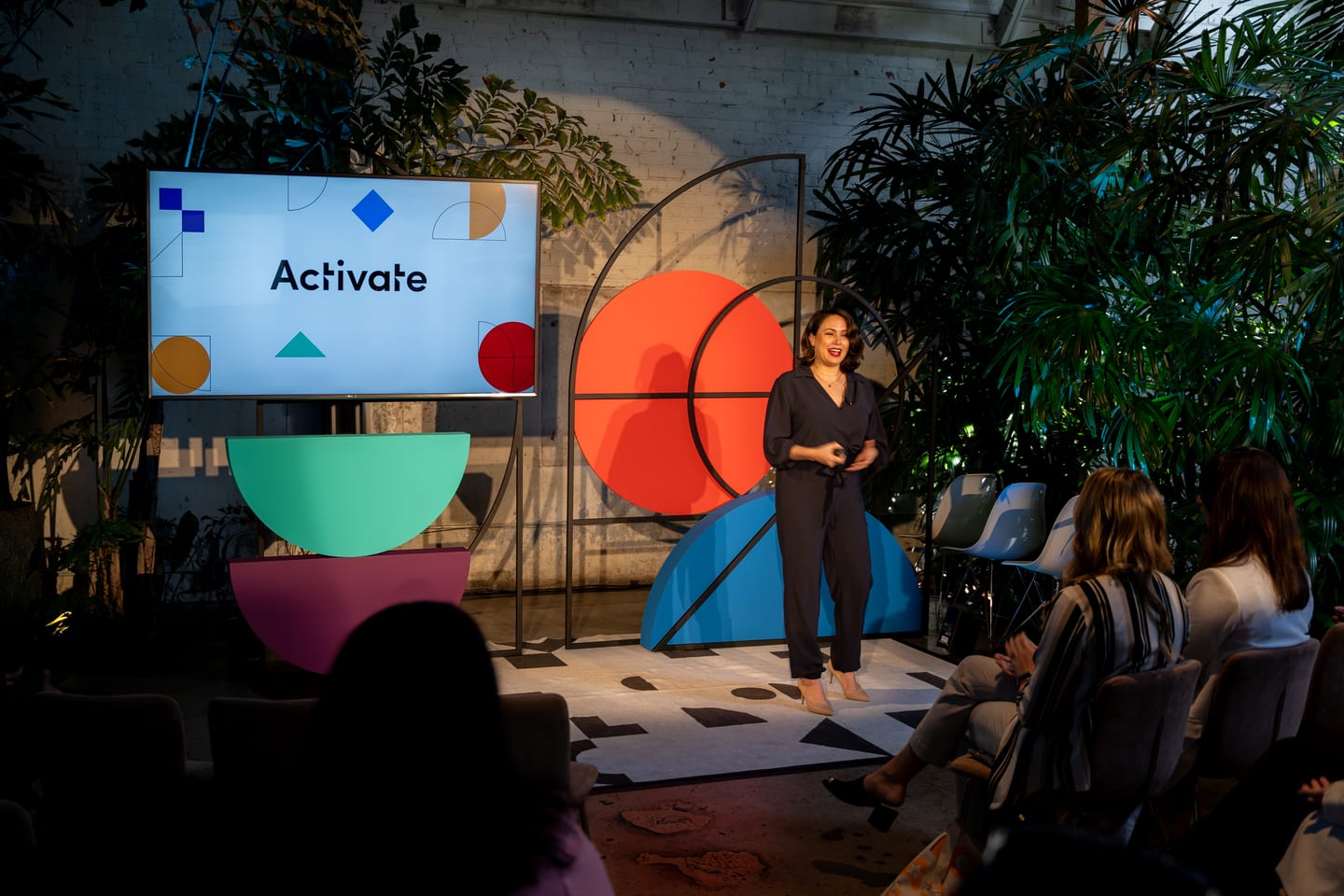 A speaker presenting beside an Activate screen on a stage of colorful geometric shapes surrounded by greenery, before an intimate audience