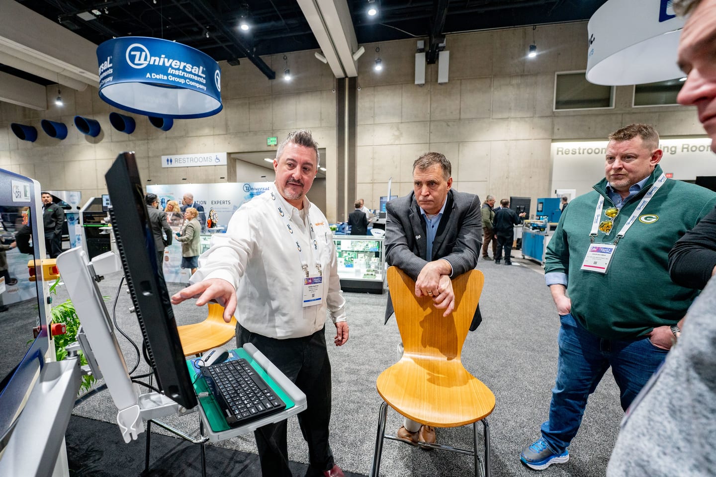 An exhibitor in an IPC-branded shirt demonstrating equipment on a monitor to two attendees on the IPC APEX EXPO show floor, a Universal Instruments banner hanging overhead