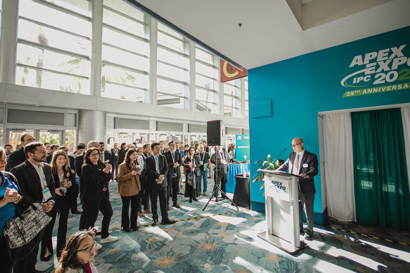 A speaker at a podium addressing a standing crowd in front of an IPC APEX EXPO 25th Anniversary wall at the Anaheim Convention Center