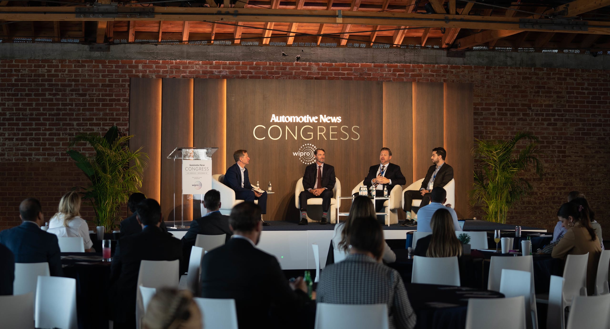 A moderator at a podium and three panelists seated on stage before a seated audience at the Automotive News Congress in Los Angeles