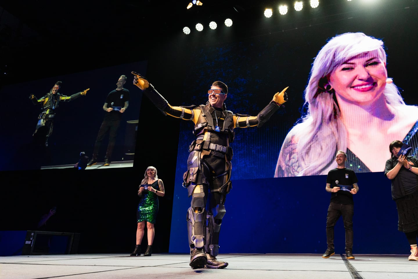 A cosplayer in a yellow-and-black armored exosuit walking the CitizenCon stage with arms spread, hosts and a big screen close-up behind him