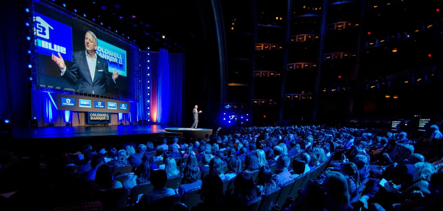 A speaker on stage before a full auditorium under blue stage light at a corporate general-session keynote