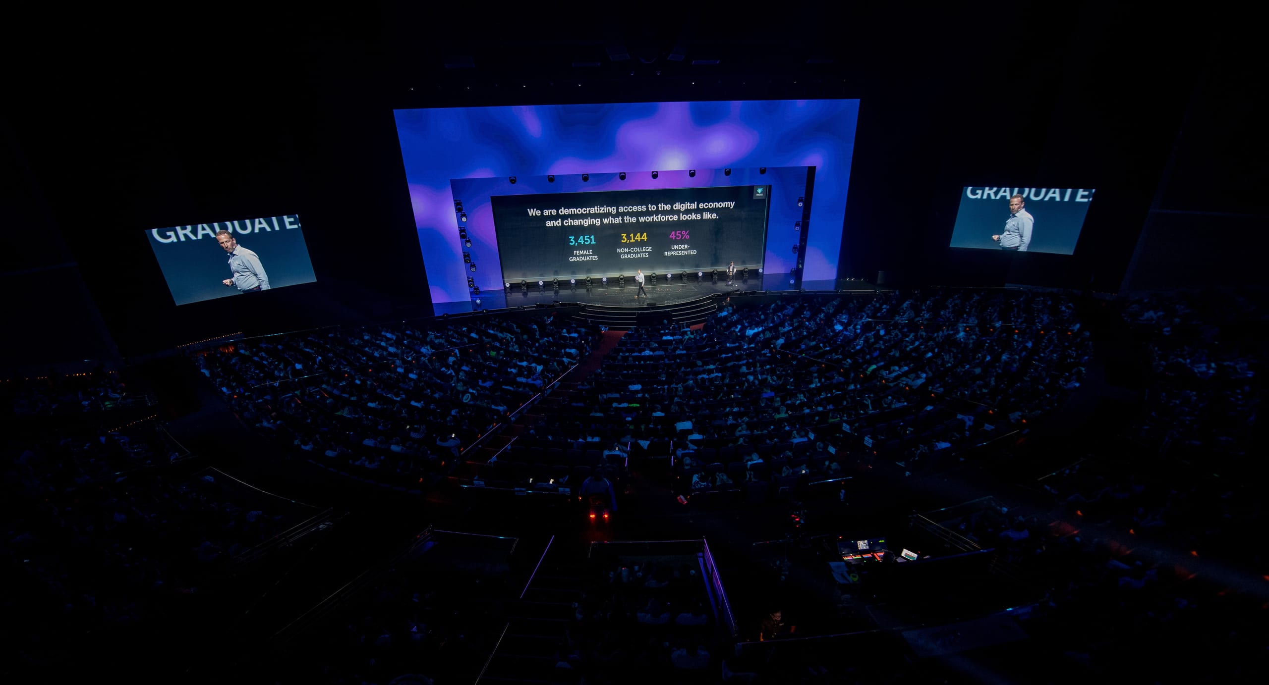 An overhead view of a packed arena keynote in Las Vegas with a triple-screen stage glowing over thousands of attendees