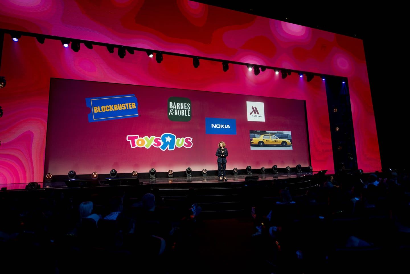A speaker on a hot-pink LED main stage in Las Vegas with Blockbuster, Barnes & Noble, Nokia, Toys“R”Us, and Marriott logos on screen