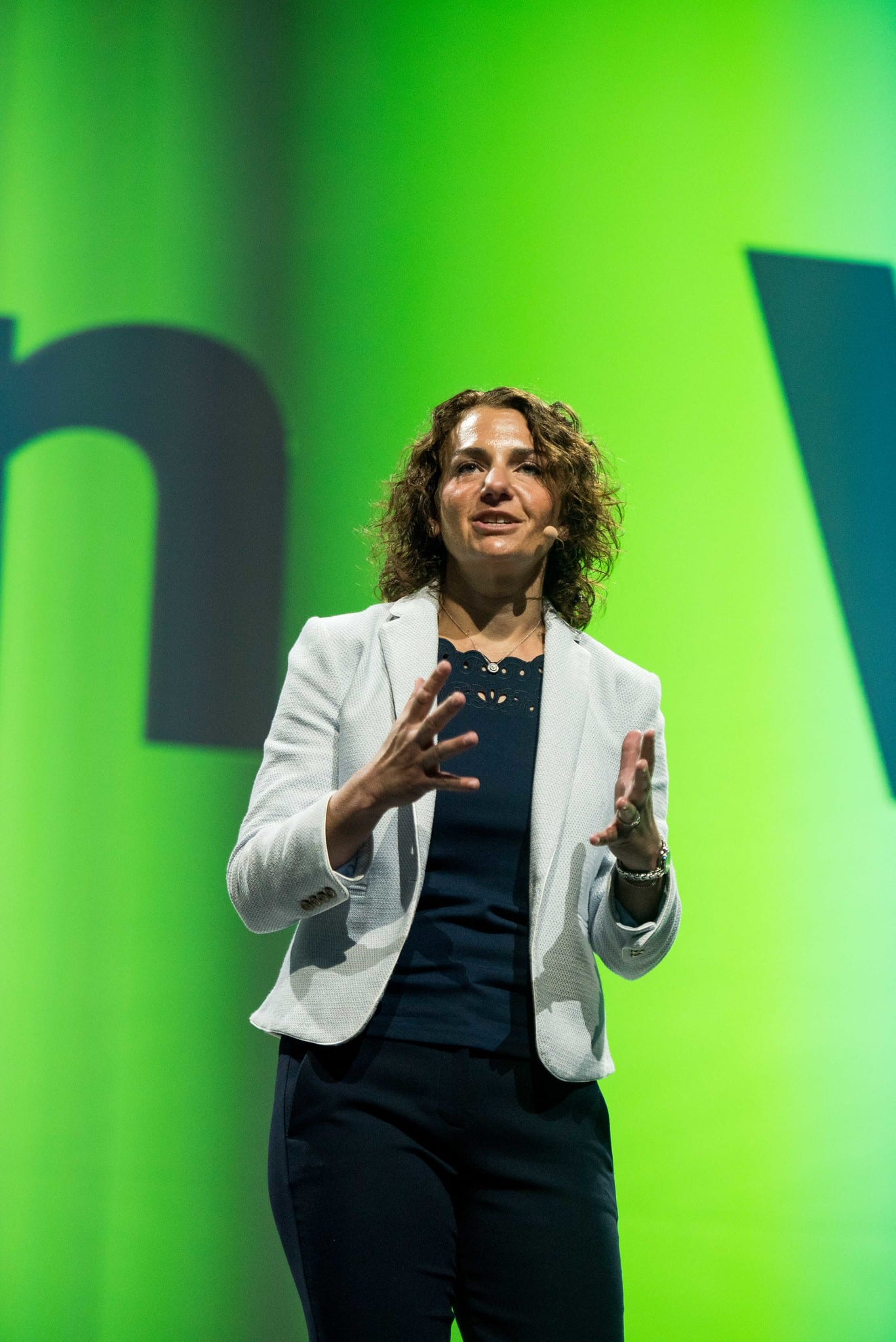 A speaker in a white blazer presenting against a bright green LED wall at a corporate keynote