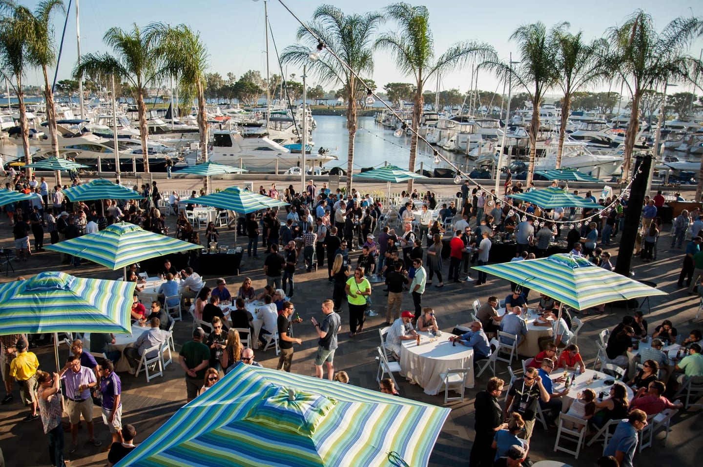 Guests dining under green striped umbrellas at an outdoor corporate reception beside a marina with sailboats and palm trees