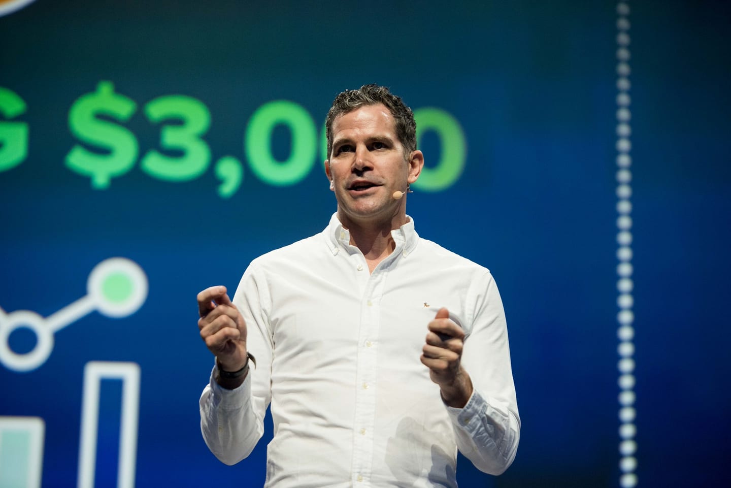 A speaker mid-presentation in front of a giant blue LED wall showing a $3,000 data chart at a corporate conference