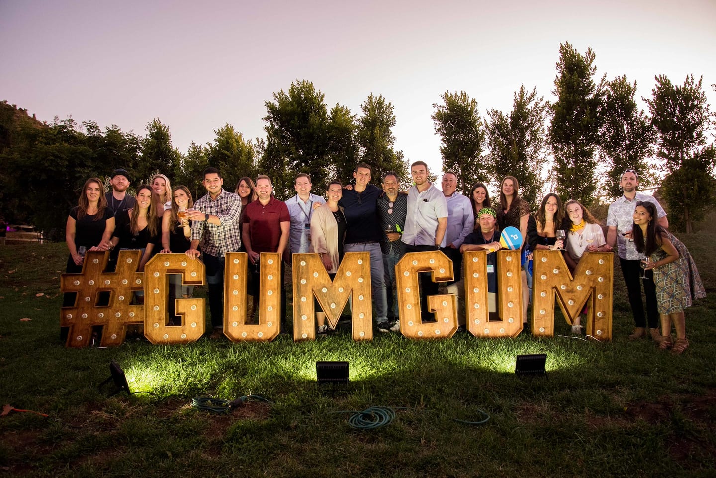 About twenty-five colleagues posed together at dusk behind giant lit marquee letters spelling hashtag GUMGUM on a grass lawn, drinks in hand
