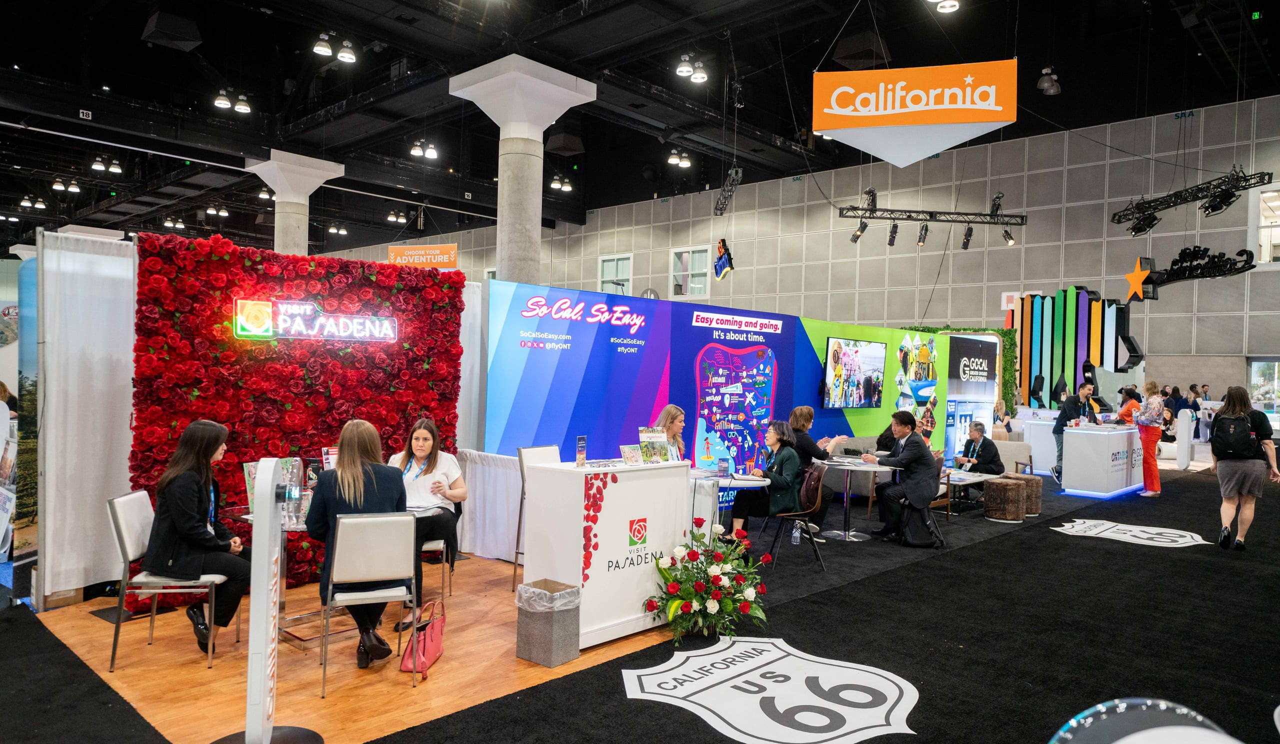A wide view of the California pavilion at the IPW travel trade show — rows of destination exhibitor booths, buyers in meetings, and a Route 66 floor graphic across the convention hall