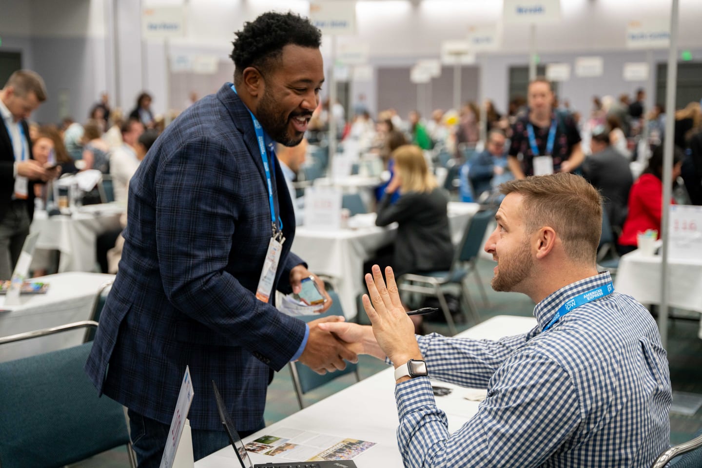 Two delegates shaking hands across an appointment table on the crowded IPW Marketplace floor at the Los Angeles Convention Center