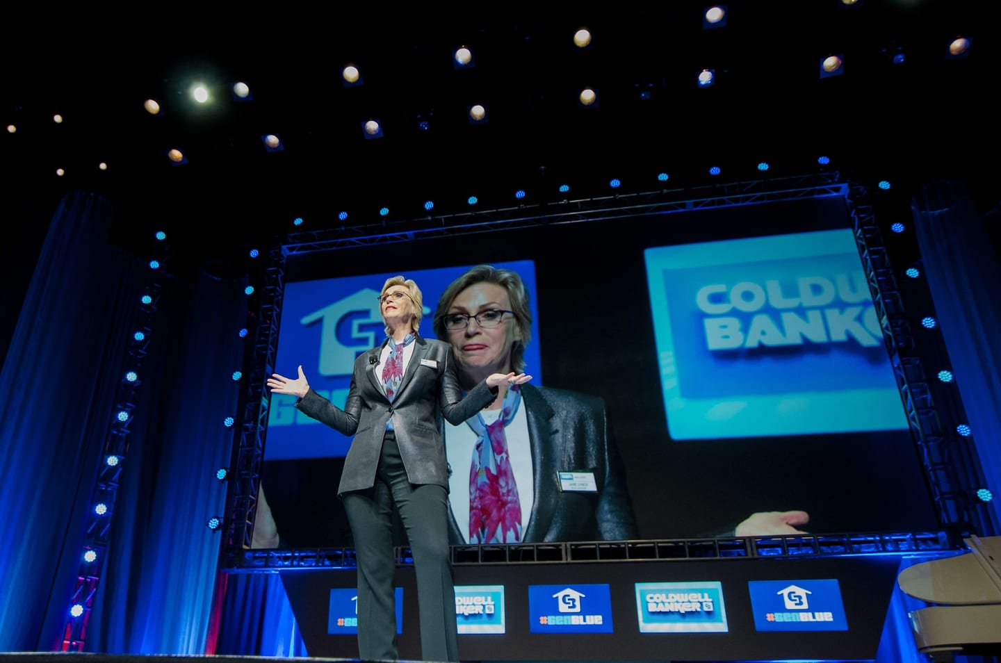 Jane Lynch hosting on the main stage in front of large screens at a Coldwell Banker event