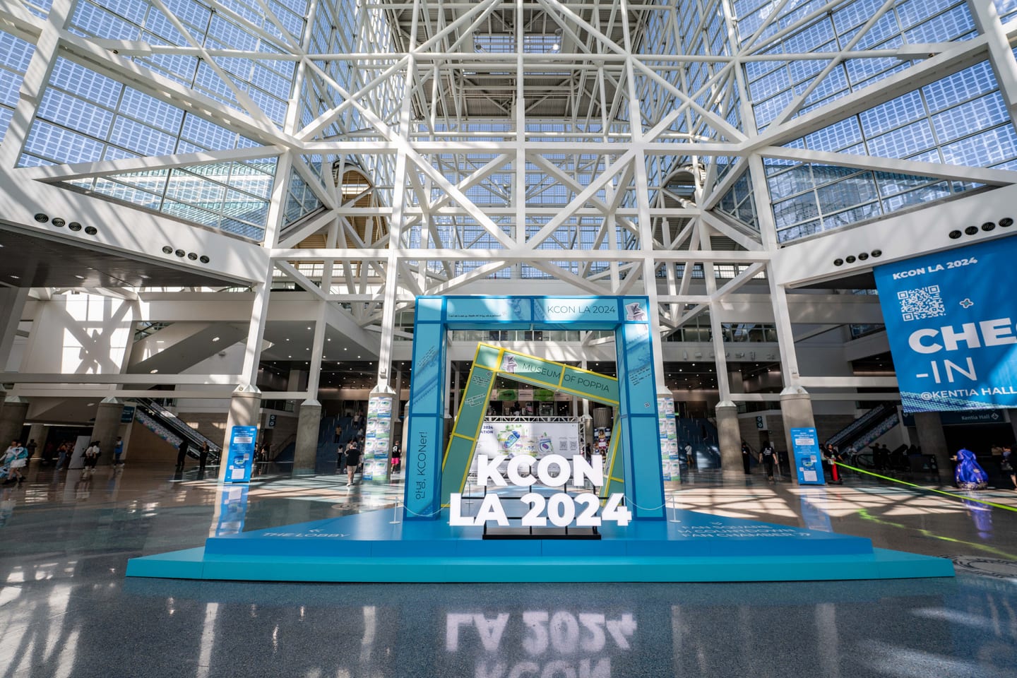 The KCON LA 2024 check-in stage under the glass atrium of the Los Angeles Convention Center, with attendees arriving across the open lobby