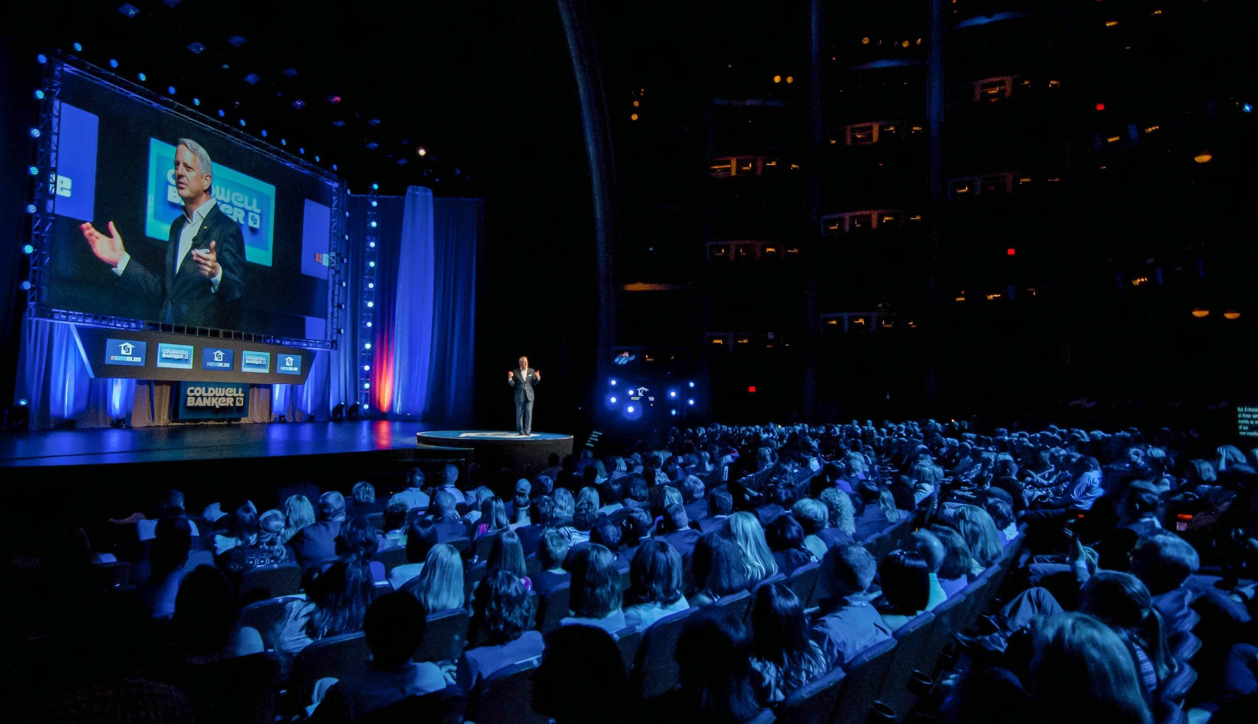 A speaker on stage under Coldwell Banker screens before a packed house, shot from the back of the house at the Dolby Theatre in Hollywood