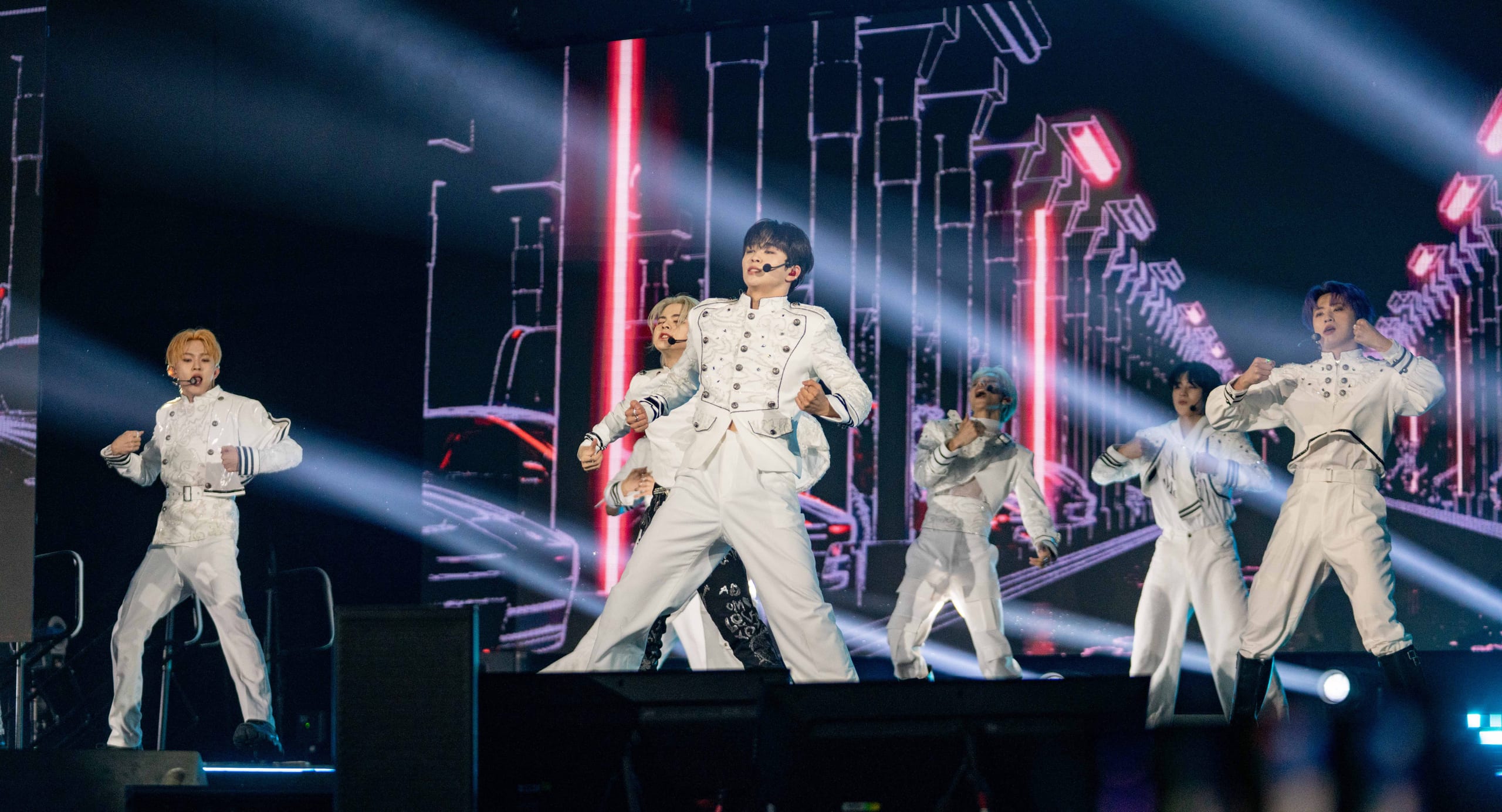 A K-pop group in white stage outfits performing under sweeping light beams on the KCON main stage with a large LED wall at the Los Angeles Convention Center