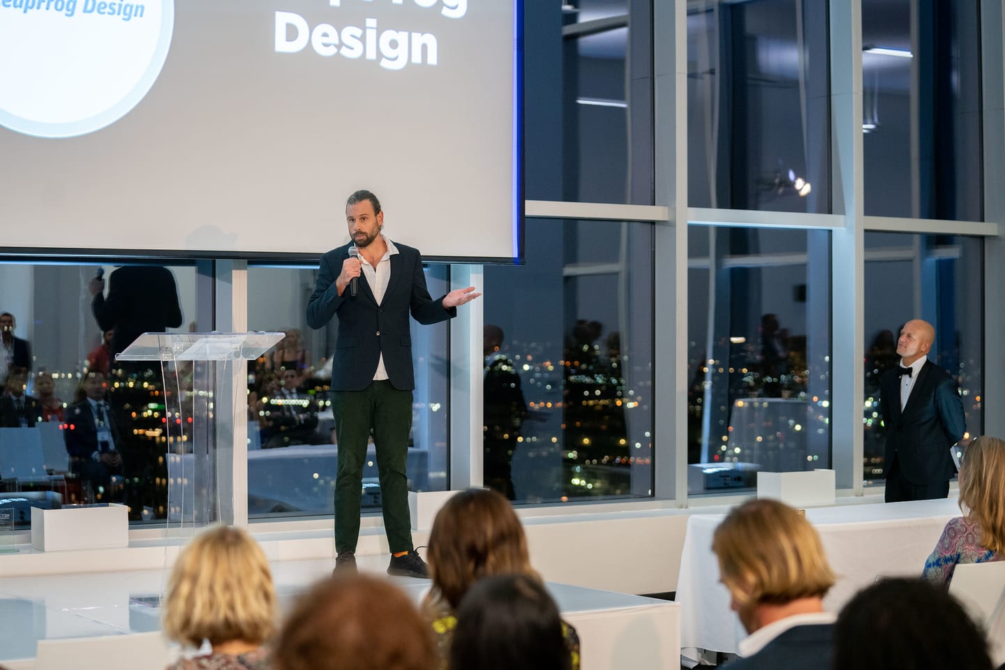 A speaker presenting beside a frog Design screen at the NetZero summit on a Downtown LA rooftop at dusk