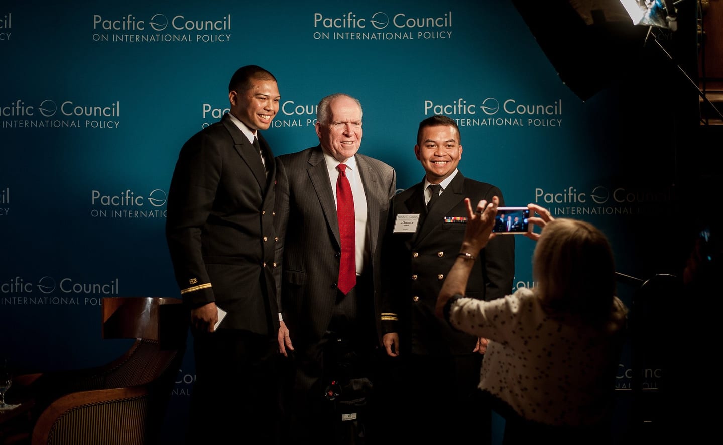 Former CIA Director John Brennan posing with two U.S. Navy officers at the Pacific Council on International Policy step-and-repeat