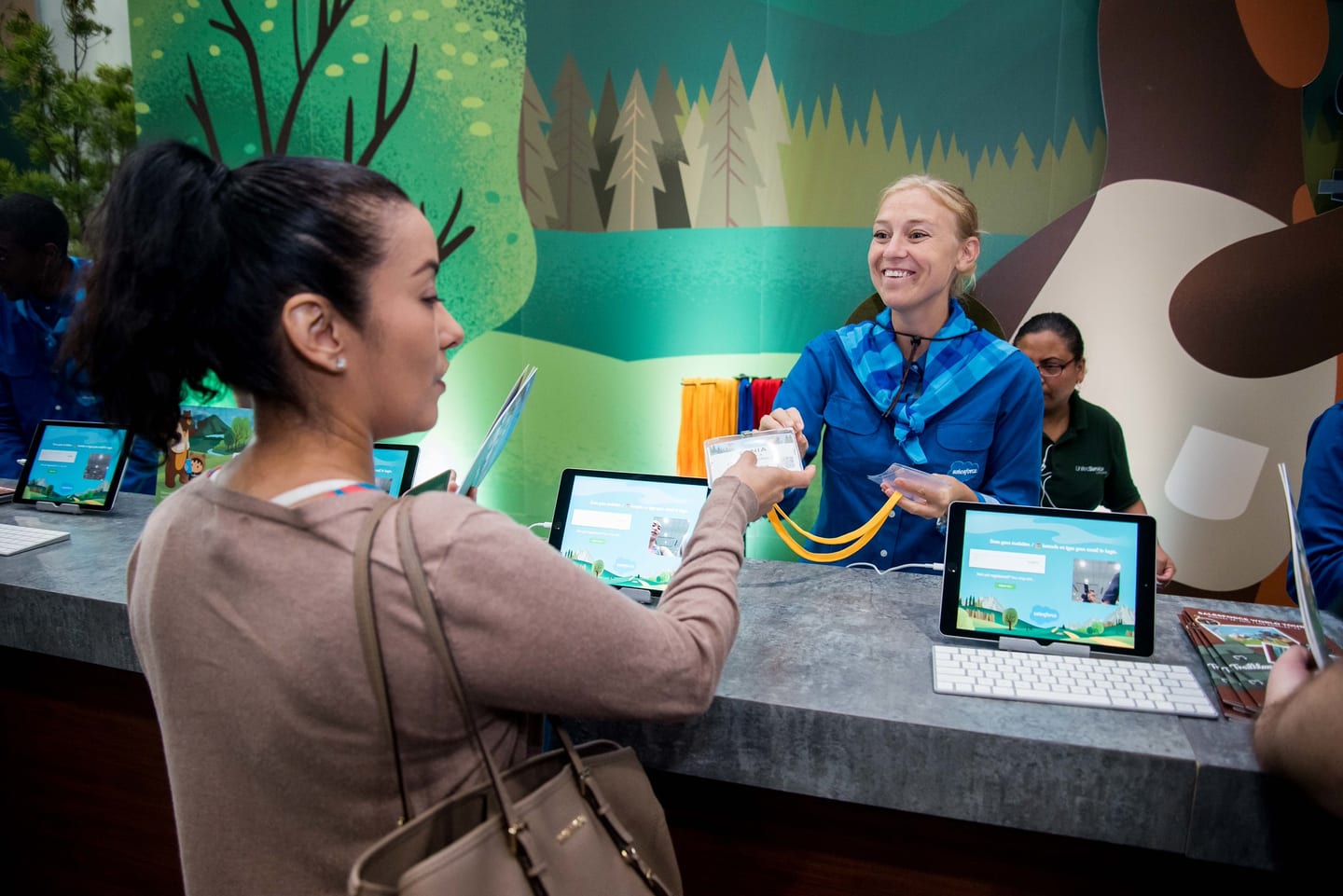 An attendee checking in at a Salesforce registration desk with an illustrated woodland backdrop and rows of tablets