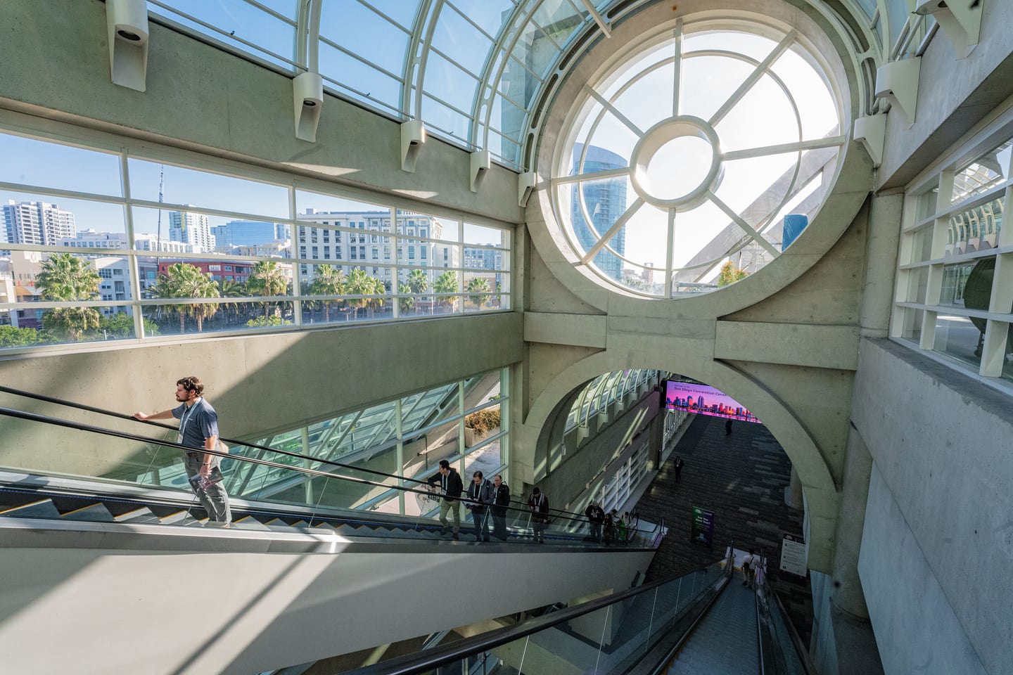 Convention attendees riding escalators beneath the circular window and digital sign inside the San Diego Convention Center
