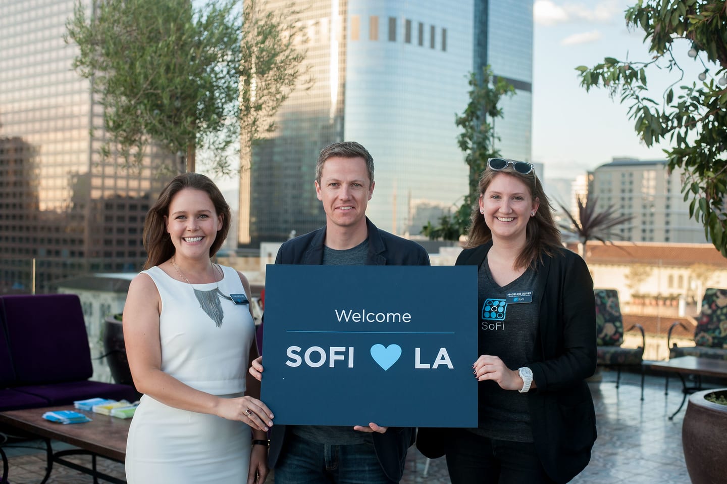 Three people holding a “Welcome — SoFi ♥ LA” sign at a rooftop reception with the Downtown Los Angeles skyline behind them