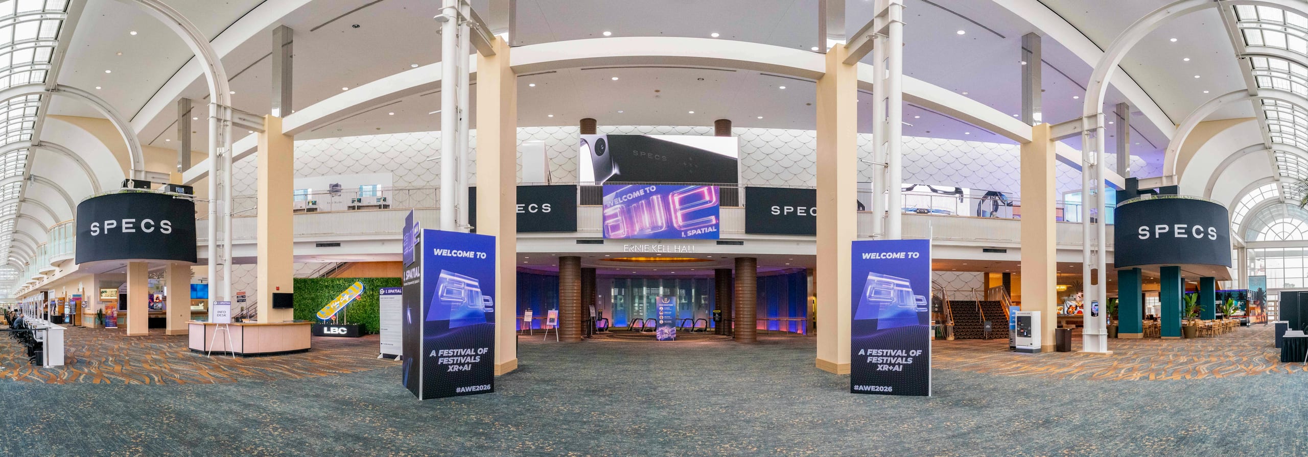 An ultra-wide view of the SPECS XR and AI festival entrance in a convention center concourse, with SPECS signage and lit welcome towers under a vaulted glass ceiling
