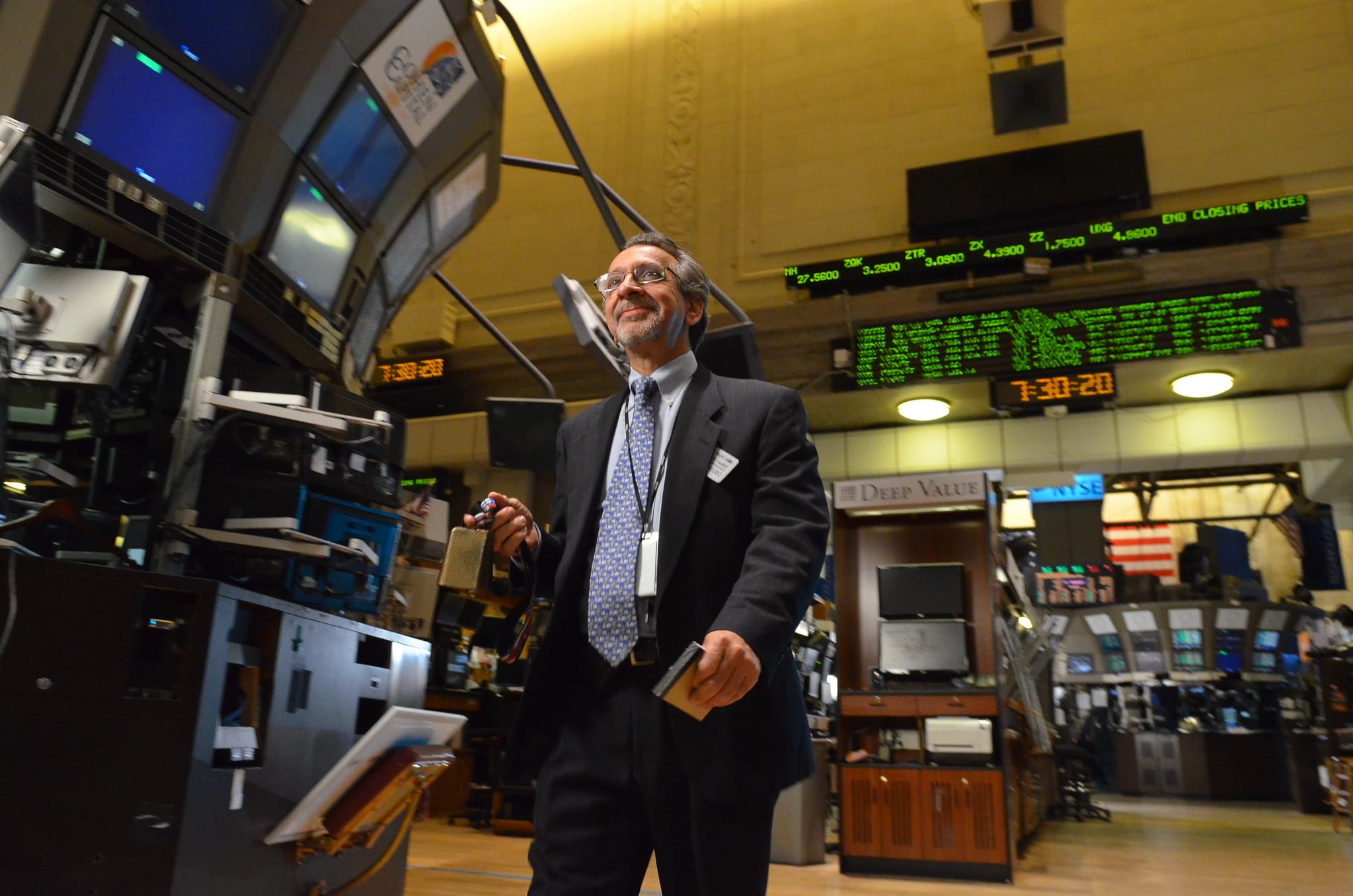 A man in a suit smiling on a Wall Street trading floor surrounded by market ticker screens in New York